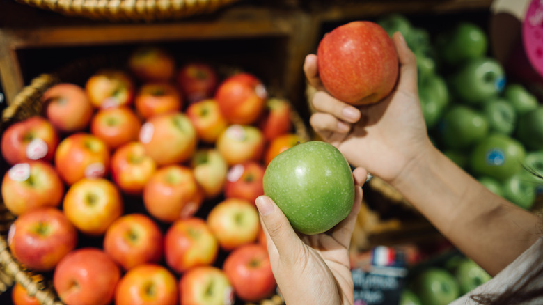 Holding a red and green apples in either hand in front of a supermarket apple display