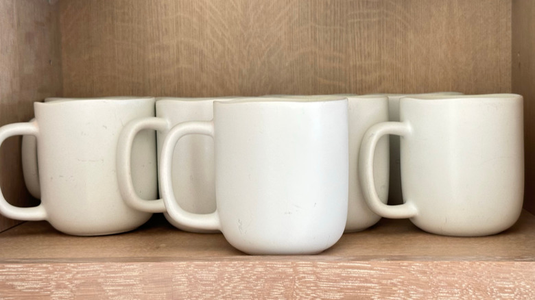 White coffee mugs lined up on the bottom shelf of a light wood cabinet
