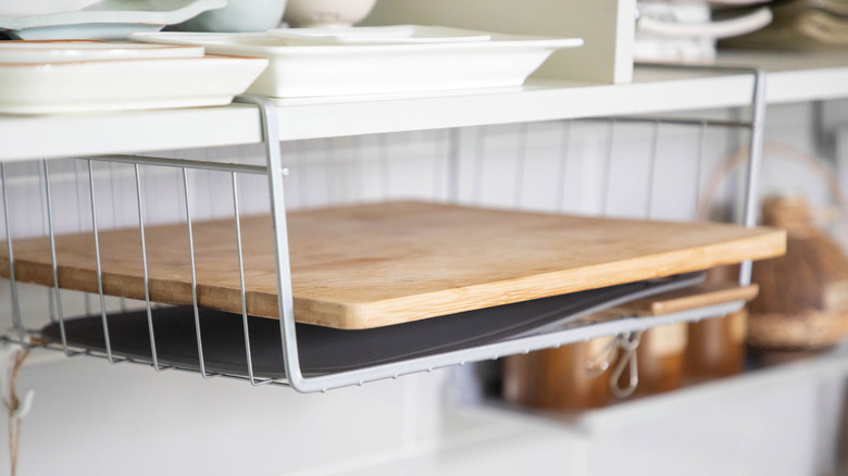 A silver wire slide-on shelf attached to the bottom of a kitchen cabinet and holding a wooden cutting board