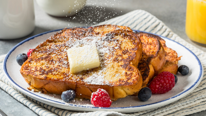 Slices of French toast on blue rimmed plate with pat of butter, raspberries, blueberries, and powdered sugar