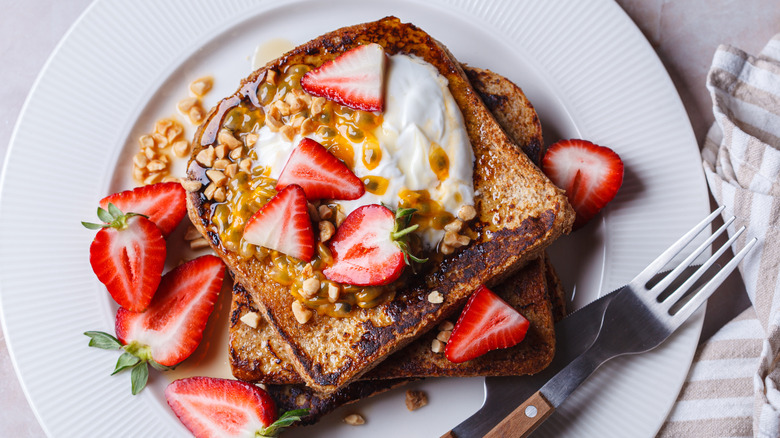 Two slices of well browned French toast on white plate garnished with cream, nuts, strawberries, and passion fruit