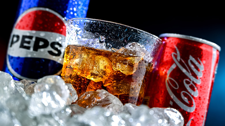 Pepsi and Coke cans on ice with glass of soda