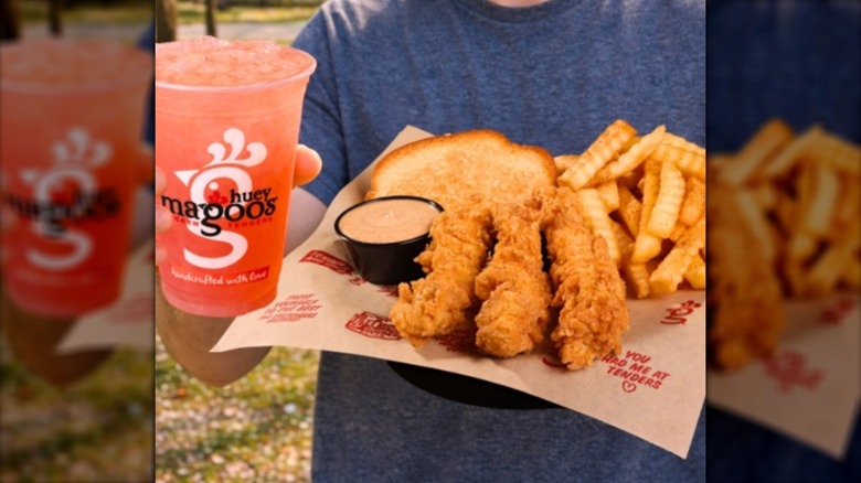 Someone holding a basket of chicken tenders, fries, and a slice of toast beside a pink drink