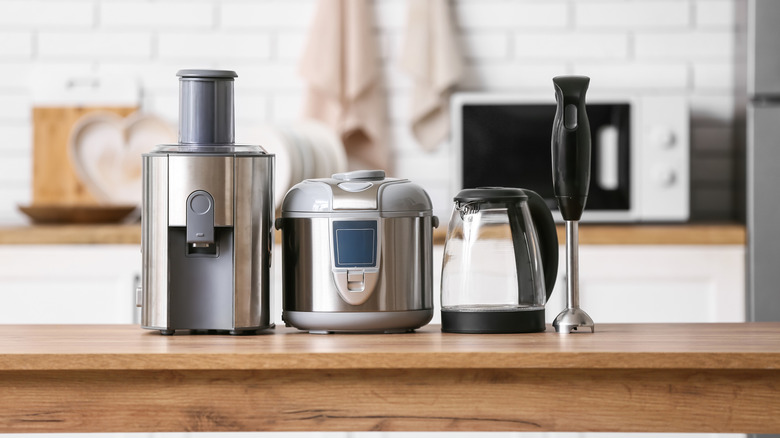 A variety of kitchen appliances including a juicer, a kettle, and an immersion blender on a countertop with dishes in a microwave in the background