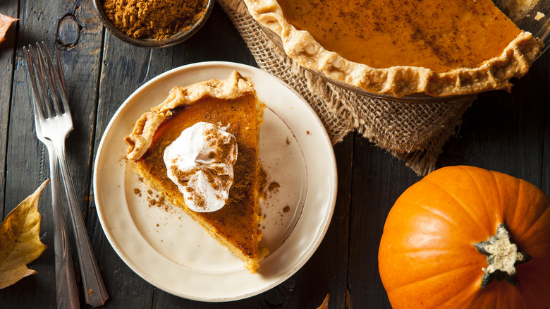 A slice of pumpkin pie on plate with the whole pie nearby, a bowl of cinnamon, forks and fall decor.