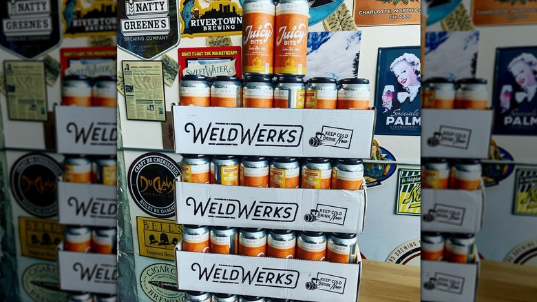 Stacked cases of Juicy Bits IPA cans in front of a poster-covered wall