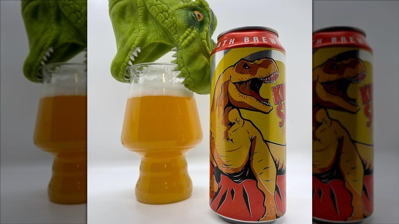 A green plastic toy dinosaur bites down on a glass of beer with a can of King Sue in the foreground