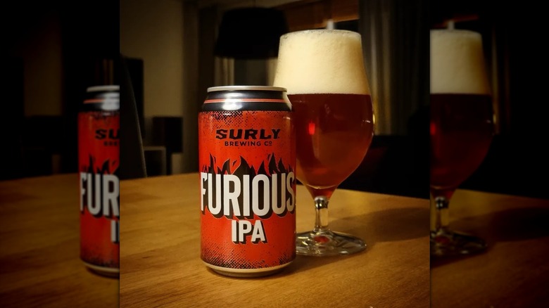 Red can of furious IPA in front of a frothy-headed glass of beer on a wooden table.