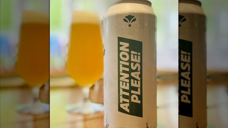 A green and white can of Attention Please! IPA in the foreground, a glass of the beer in the background.