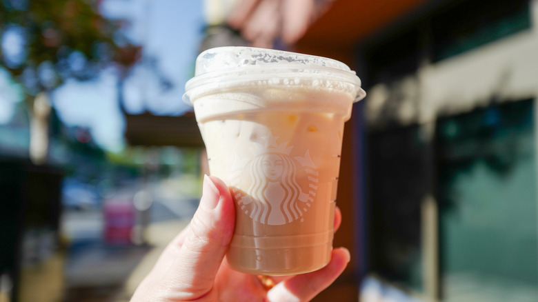 Someone holds up an iced chai latte from Starbucks outside