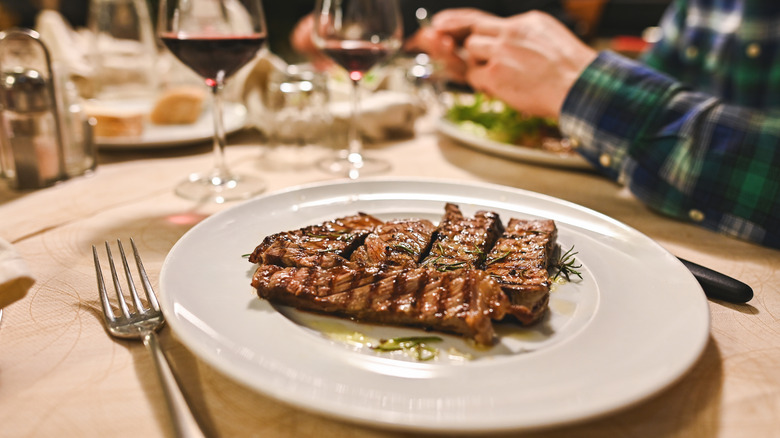 A portion of grilled steak on a plate with wine and utensils around it