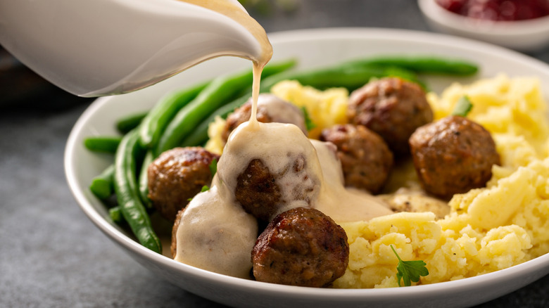 A person pouring creamy gravy over a plate of Swedish meatballs, mashed potatoes, and green beans