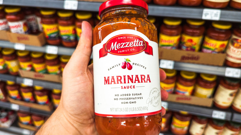 A hand holds a jar of Mezzetta Marinara in a grocery store aisle.