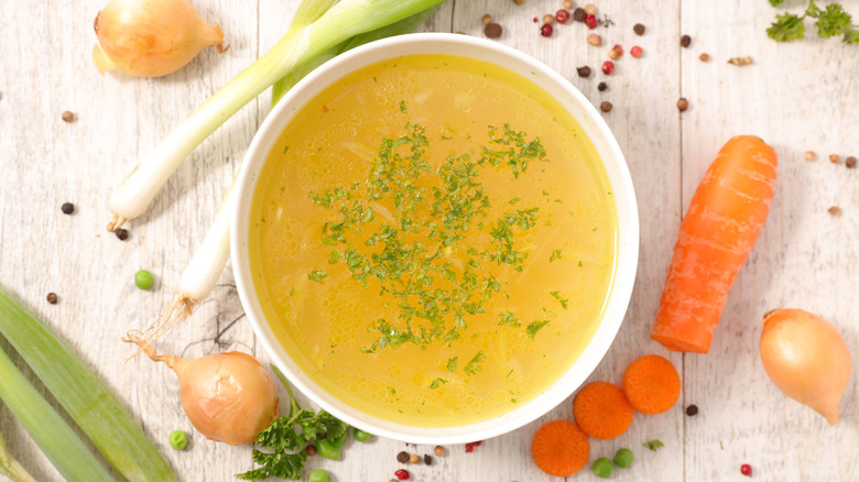 A white bowl of vegetable broth garnished with chopped parsley surrounded by raw vegetables