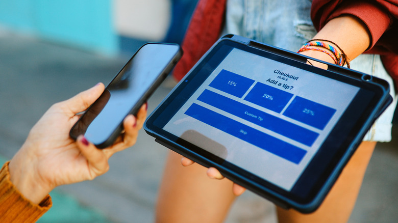 Person holding a phone next to a large tipping screen with 15%, 20%, and 25% displayed on a tablet