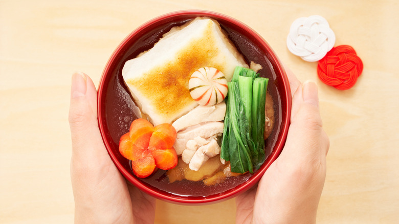 Hands holding red bowl of Japanese ozoni soup with grilled mochi cake, carrots, chicken, and greens