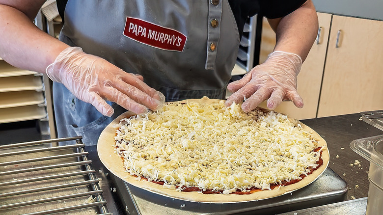 A Papa Murphy's employee putting shredded cheese on a pizza
