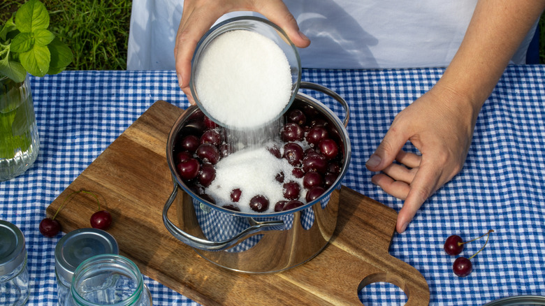 A person pours sugar into a pan of cherries on a gingham tablecloth