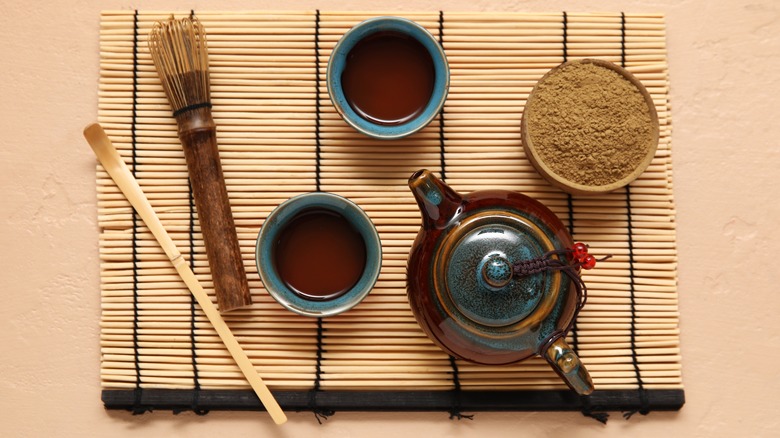 Overview of a bamboo mat with a bowl of hojicha powder, a bamboo whisk, a brown teapot, and two cups of tea