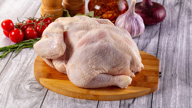 A large, raw chicken sitting on a wooden chopping board, with cherry tomatoes on the vine, a garlic bulb, a red onion, and salt and pepper shakers in the background