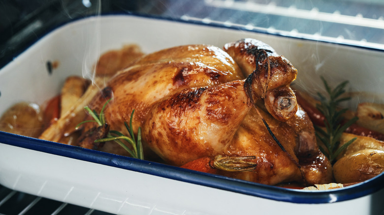 A roast chicken in a roasting pan, surrounded by root vegetables and herbs, resting in an oven