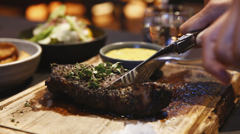 person slicing herb-topped steak on a wooden cutting board