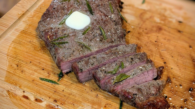 a sliced steak topped with rosemary and butter on a wooden cutting board