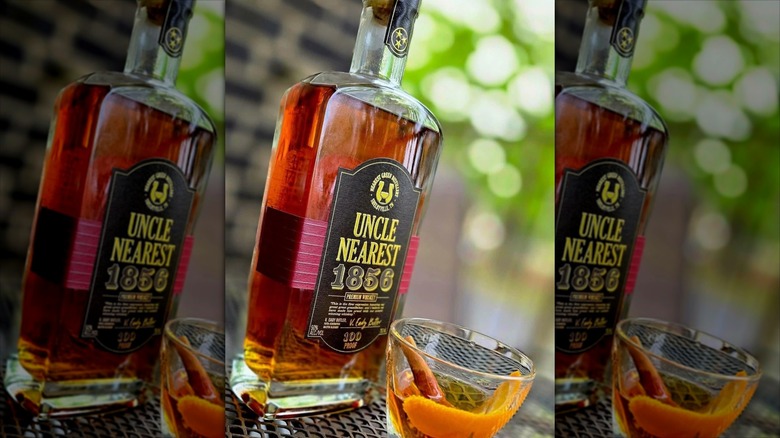 A bottle of Uncle Nearest whiskey on a table next to a glass with an orange rind and cinnamon stick