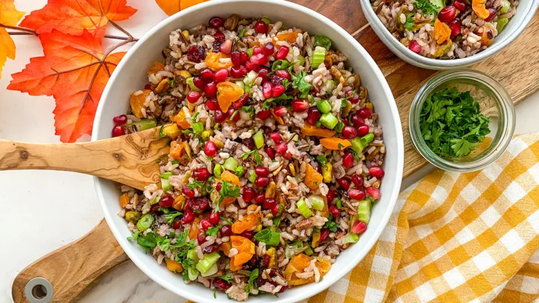 Orange and yellow leaves on a white marble table are topped with a wooden cutting board and yellow plaid napkin. On top of the cutting board are two white bowls, one large, one small, filled with a wild rice pilaf that has wild rice, sweet potatoes, cranberries, celery, green onions, and nuts. There is a small glass bowl with extra chopped cilantro in between the two bowls of wild rice pilaf