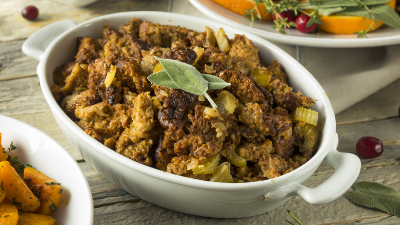 A white serving baking dish is filled with stuffing and topped with two pieces of sage leaves. The photo shows some of the ingredients in the stuffing including chopped celery and walnuts.