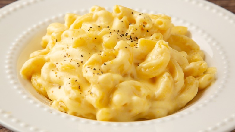 A white serving bowl plate is filled with a yellow creamy macaroni and cheese, with a sprinkle of pepper and other seasonings on top