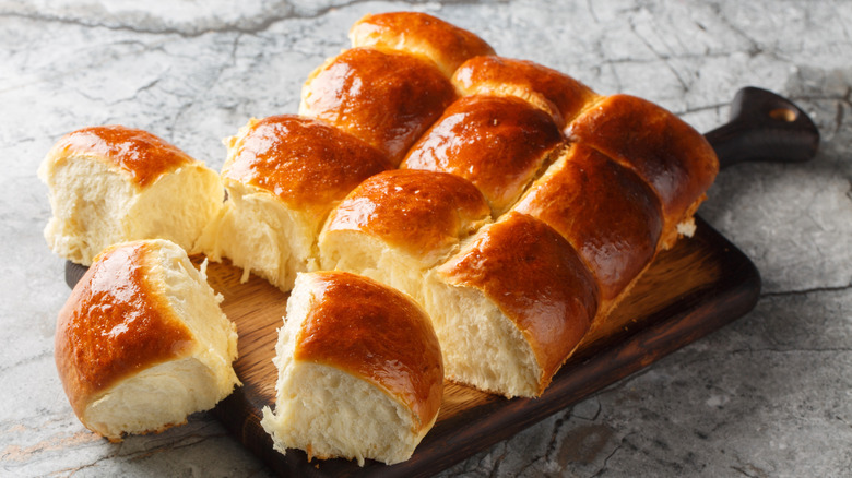 Twelve dinner rolls glistening on top are placed on a wooden cutting serving board on top of a dark gray marble counter. Three dinner rolls in front are detached from the rest.
