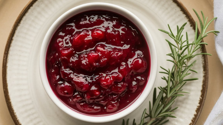 A rosemary sprig is on the right side of a white dinner plate, and topped with a bowl filled with homemade cranberry sauce ruby red in color, showing cranberry lumps.