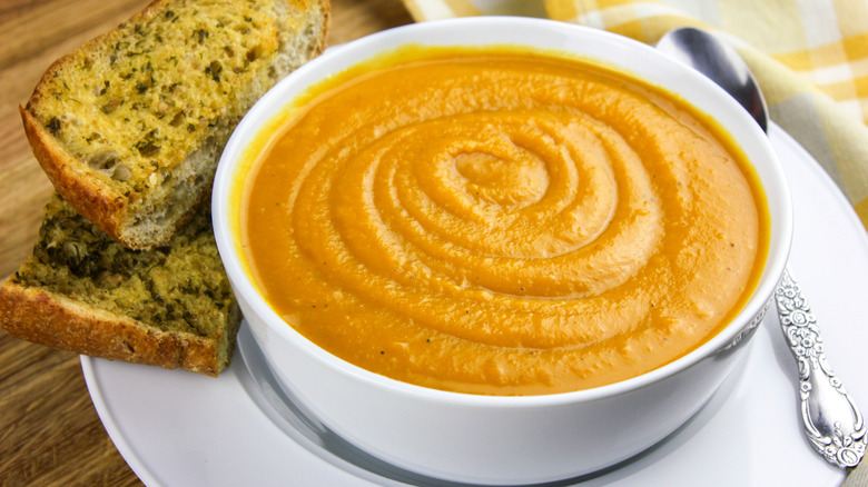 Toasted garlic bread cut in half and stacked on top of one another is on a white plate next to a white bowl filled to the brim with a yellow-orange butternut squash soup. A silver spoon rests on the plate on the opposite side of the garlic bread, next to a yellow plaid napkin.