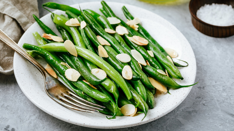A white dinner plate is topped with blanched green beans topped with slivered almonds. A fork is on the plate next to a linen beige napkin, and  a small dark brown ramekin of salt