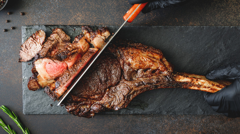 Black-gloved hands slicing a dry-aged tomahawk steak on a slate board.