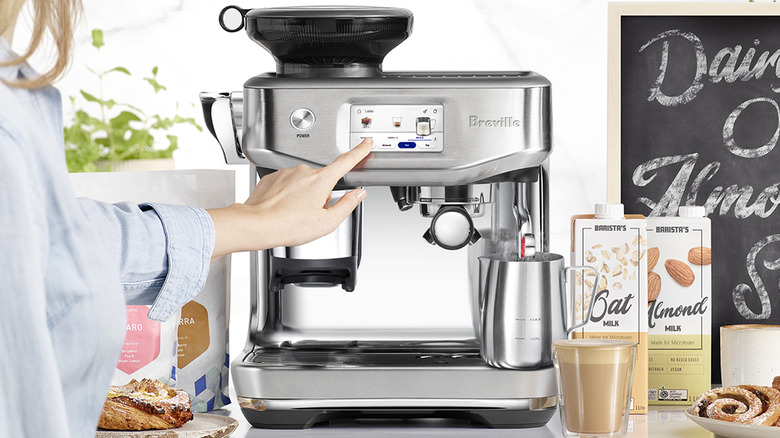 Someone touches the screen of the Breville Barista Touch Impress on a counter surrounded by milks and coffees