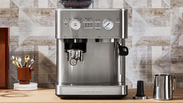 The KitchenAid Semi Automatic Espresso Machine with Burr Grinder on a counter next to a jug, tamper, and bowl of spoons