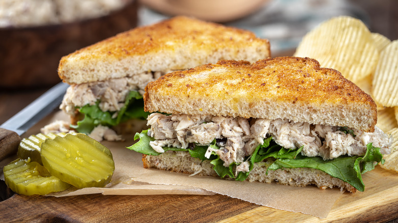 a tuna salad sandwich with lettuce on a wooden cutting board beside pickles and potato chips
