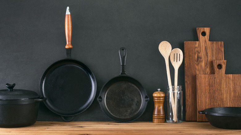 Two cast iron pans, a cast iron dutch oven, two wooden cutting boards, and wooden utensils on a wooden counter with a black background
