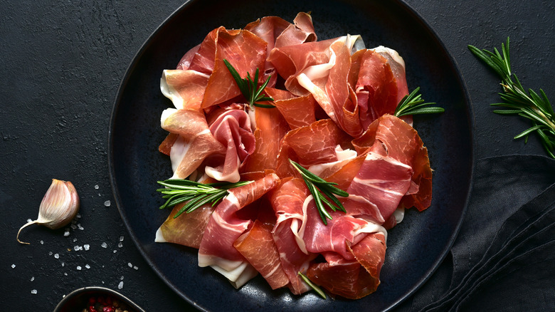 A black plate with slices of prosciuuto and iberico hams, rosemary and garlic garnish