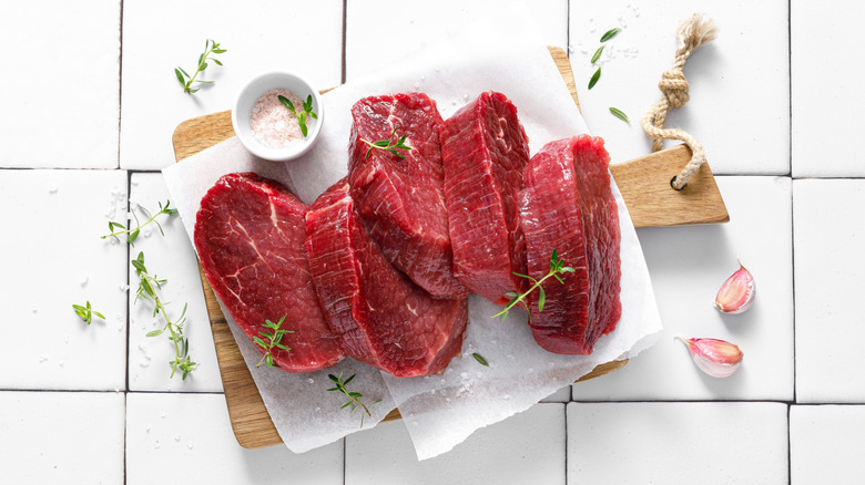raw beef tenderloin steaks on parchment paper on a wooden cutting board with garlic, salt and herbs nearby and a white tile background.