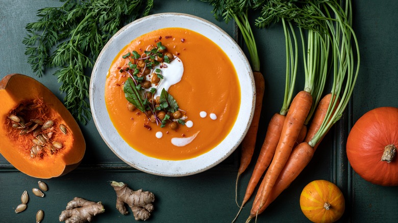 A white bowl filled with a creamy orange pumpkin chickpea chili is topped with microgreens, chickpeas, and cream, on a dark green table, surrounded by fresh ingredients included in the stew such as pumpkin, ginger, carrots, and butternut squash