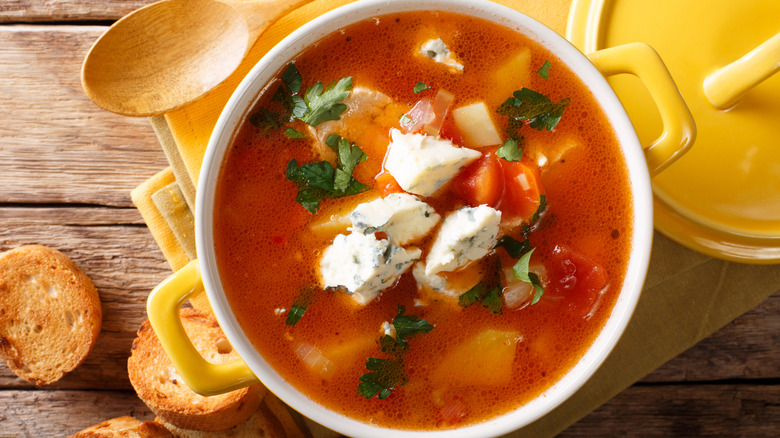 A light wooden table is topped with a closeup of a yellow bowl of buffalo chicken soup with clumps of bleu cheese and fresh cilantro on top, toasted slices of baguette are scattered on the table