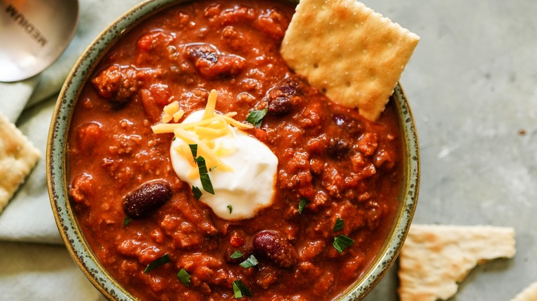 Traditional beef chili served in a green bowl and topped with shredded cheese, sour cream, and served with saltine crackers