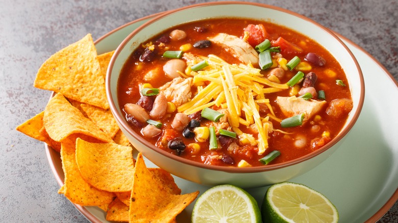 A bowl of soup with sweet corn and black bean soup amongst other visible vegetables is topped with shredded cheddar cheese and served on a matching plate with tortilla chips and lime cut in half