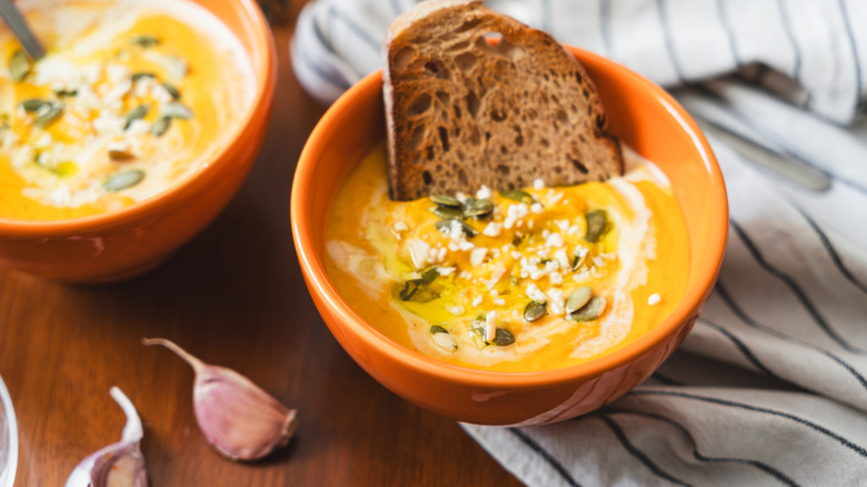 Two orange bowls are filled with a creamy orange-yellow butternut squash soup topped with pumpkin seeds, and a slice of whole wheat toast served on a wooden table