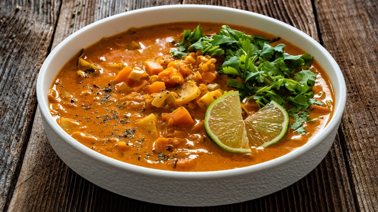 A wooden table is topped with a white bowl filled with an orange-colored creamy stew made with coconut milk and sweet potatoes. Seasoning, including pepper, is visible on the top layer of the stew, along with a pile of fresh cilantro, and two thin-sliced lime wedges