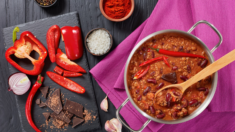 A dark wooden table is topped with a cutting board featuring sliced onion, sliced red pepper, and a red chili, along with a bowl of salt, peppercorns, and chili powder, next to a vibrant purple tablecloth topped with a metal pot of chili made with the ingredients reflected on the table