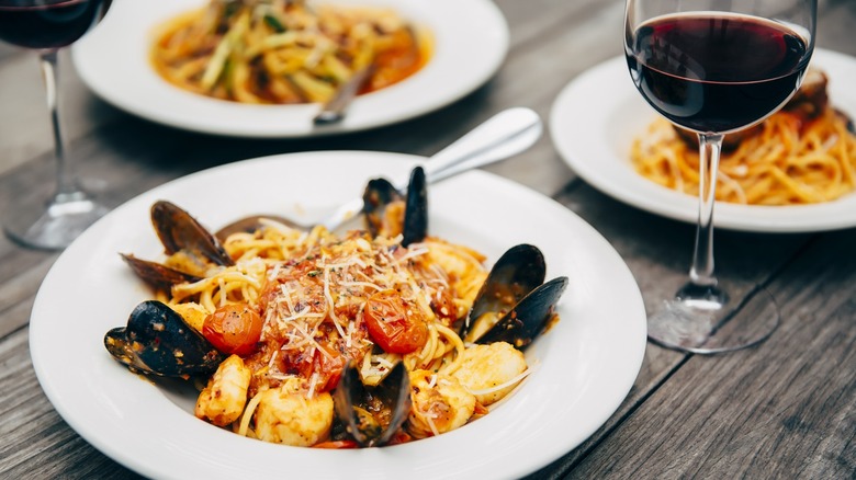 A bowl of pasta with mussels and tomatoes and cheese on a wooden table with a glass of wine
